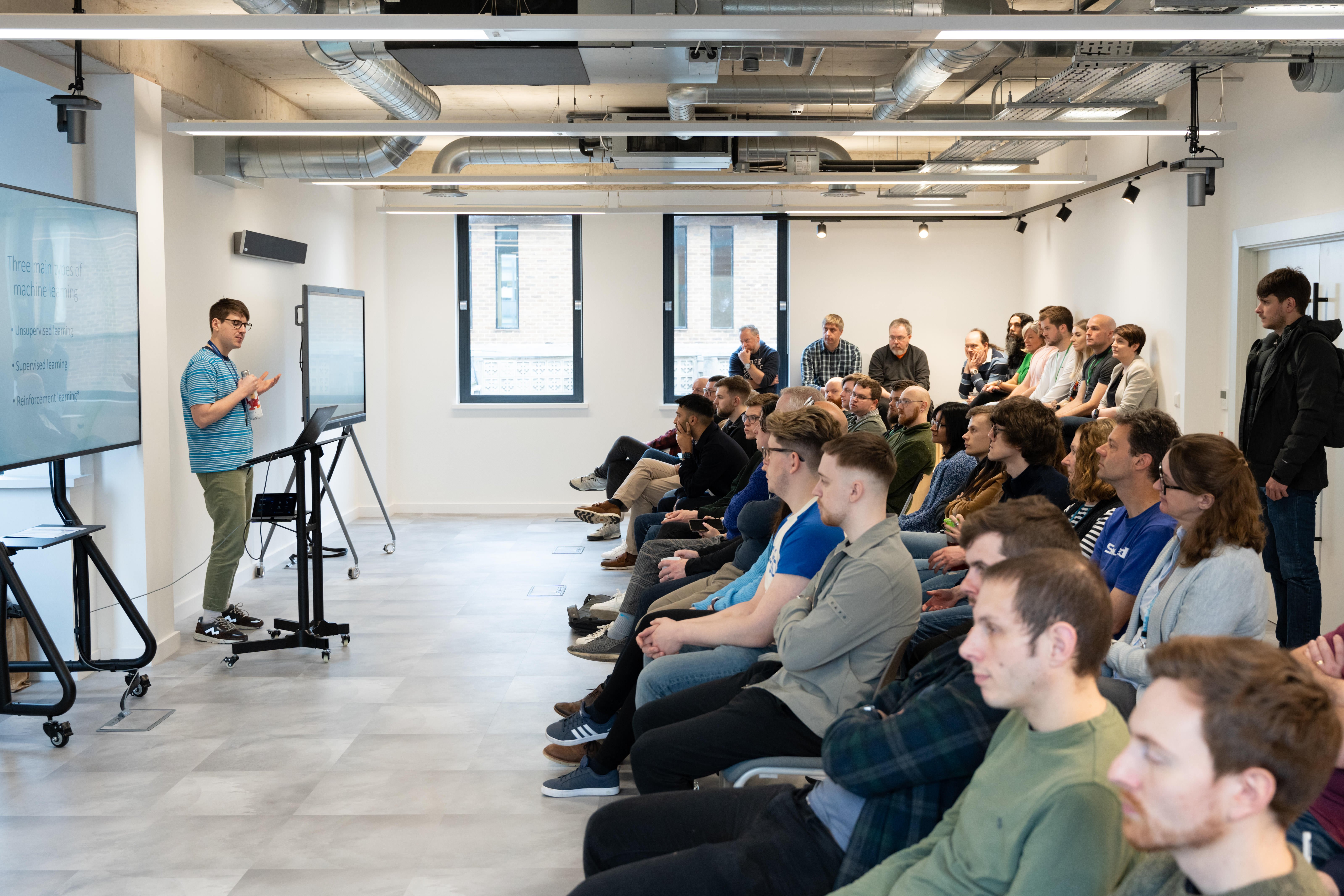 Image of person standing at front of room, with other people in front of them, seated in rows.